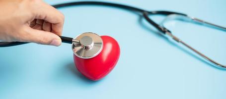 main de médecin tenant un stéthoscope avec une forme de coeur rouge sur fond bleu. soins de santé, assurance-vie et concept de la journée mondiale du cœur