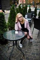 fille blonde à lunettes et manteau rose, tunique noire assis à table café en plein air. photo