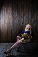 fille blonde élégante portant un manteau de fourrure assis sur une chaise au studio sur fond de bois. photo