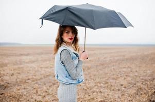 portrait de jeune fille bouclée brune en veste jeans avec parapluie noir sur le terrain. photo