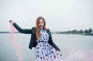 une fille élégante en veste de cuir tient une fusée de fumée rose le jour de l'hiver contre un lac gelé. photo