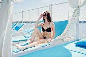 portrait d'une jolie brune en bikini noir assis sur le transat avec des lunettes de soleil au bord du lac. photo