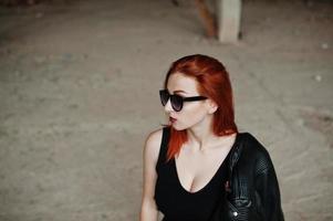 fille élégante aux cheveux rouges portant des lunettes de soleil en noir, contre un endroit abandonné. photo