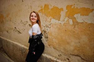 élégante fille blonde porter sur une veste en cuir noir posant dans les rues de la ville fond vieux mur avec des ombres. photo