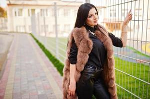 photo de mode en plein air d'une magnifique femme sensuelle aux cheveux noirs dans des vêtements élégants et un luxueux manteau de fourrure sans manches contre une clôture de fer à la ville d'automne.
