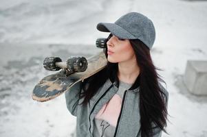 fille brune élégante en casquette grise, style de rue décontracté avec planche à roulettes le jour de l'hiver. photo