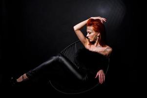 modèle de mode fille aux cheveux rouges avec à l'origine maquillage comme prédateur de léopard isolé sur noir. portrait en studio sur chaise. photo