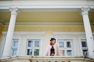 fille aux cheveux rouges dans des verres et un chapeau avec un sac à main rouge posé près de la maison vintage. photo