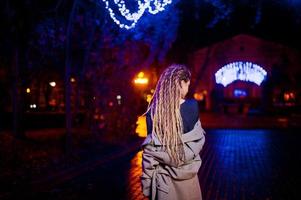 fille avec des dreadlocks marchant dans la rue de nuit de la ville contre les lumières de la guirlande. photo