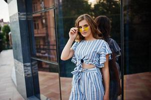 portrait d'une jeune femme parfaite portant des lunettes de soleil rayées et jaunes pose sur un balcon d'un bâtiment moderne dans une ville. photo