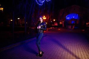 portrait de nuit du modèle de fille portant des lunettes, des jeans et une veste en cuir contre la guirlande de lumières bleues de la rue de la ville. photo