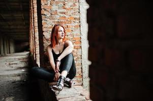 fille élégante aux cheveux rouges portant des lunettes de soleil en noir, contre un endroit abandonné avec des murs de briques. photo