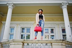 fille aux cheveux rouges dans des verres et un chapeau avec un sac à main rouge posé près de la maison vintage. photo