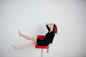 fille aux cheveux rouges sur une tunique de robe noire assise sur une chaise rouge contre un mur blanc dans une salle vide. photo