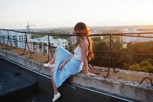 portrait d'une superbe jeune femme en t-shirt blanc et jupe bleue assise sur le bord de l'immeuble au coucher du soleil. photo