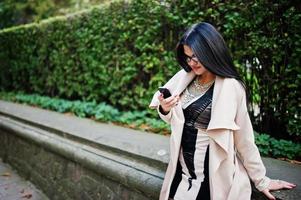 femme sexy aux cheveux noirs dans des verres et un manteau posés dans la rue avec un téléphone portable. photo