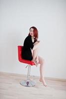 fille aux cheveux rouges sur une tunique de robe noire assise sur une chaise rouge contre un mur blanc dans une salle vide. photo