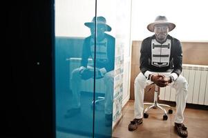 riche homme africain assis sur une chaise dans la chambre. portrait d'un homme noir réussi au chapeau à l'intérieur. photo