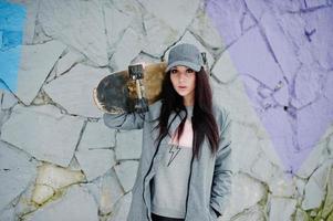 fille brune élégante en casquette grise, style de rue décontracté avec planche à roulettes le jour de l'hiver contre un mur coloré. photo