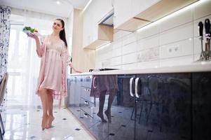 jolie fille en robe rose posant avec une assiette de salade dans la cuisine. photo