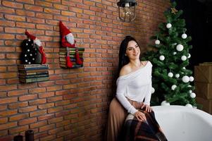 fille brune en robe posée près de l'arbre du nouvel an avec décoration de noël dans la salle de studio en brique. photo