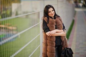 photo de mode en plein air d'une magnifique femme sensuelle aux cheveux noirs dans des vêtements élégants et un manteau de fourrure luxueux et avec un sac à dos contre une clôture en fer.