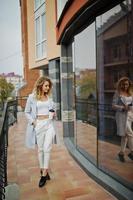 fille modèle blonde bouclée élégante porter sur blanc avec une tasse de café à portée de main posant contre une grande fenêtre. photo