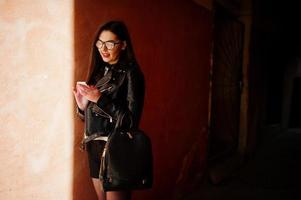 fille brune élégante porte une veste en cuir et un short avec sac à dos et lunettes contre un mur orange à l'ombre regardant sur un téléphone portable. photo