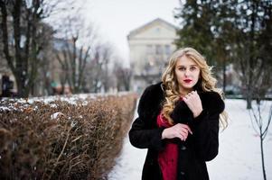 élégance fille blonde en robe de soirée rouge et manteau de fourrure dans les rues de la ville en journée d'hiver. photo