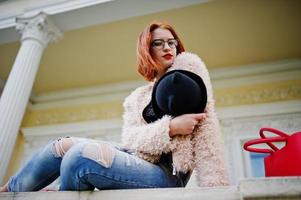 fille aux cheveux rouges dans des verres et un chapeau avec un sac à main rouge posé près de la maison vintage. photo