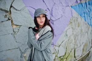 fille brune élégante en casquette grise, style de rue décontracté le jour de l'hiver contre un mur coloré. photo