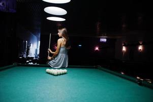 portrait d'une jolie jeune femme en robe assise sur la table de billard et tenant une queue. photo