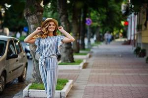 portrait d'une très jolie jeune femme en salopette rayée posant avec son chapeau sur un trottoir dans une ville avec des arbres en arrière-plan. photo