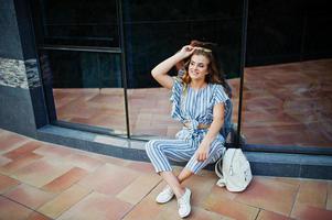 portrait d'une fabuleuse jeune femme en combinaison rayée et lunettes de soleil assise sur le sol et posant avec un sac à dos contre un mur de verre. photo