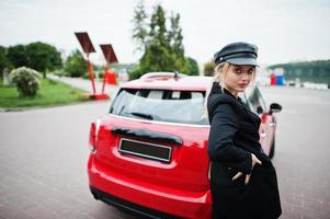 portrait d'une belle mannequin blonde sexy en casquette et en noir avec un maquillage lumineux près d'une voiture de ville rouge. photo