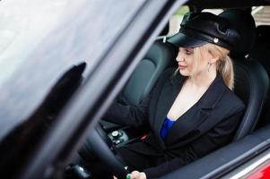 portrait d'une belle mannequin blonde sexy en casquette et en noir avec un maquillage lumineux s'asseoir et conduire une voiture de ville rouge. photo