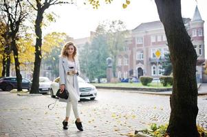 fille modèle blonde bouclée élégante porter sur blanc avec une tasse de café à portée de main posant à la rue d'automne de la ville contre la voiture blanche. photo