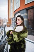 fille brune en manteau de fourrure vert dans la rue de la ville contre la maison avec de grandes fenêtres en hiver. photo