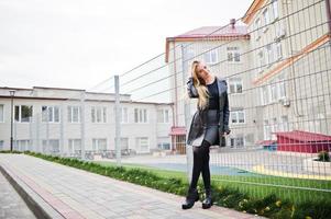 fille blonde à la mode en long manteau de cuir noir contre une clôture de fer dans la rue. photo