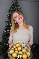 jolie fille blonde en robe beige contre l'arbre du nouvel an avec des boules dorées du nouvel an au studio. joyeuses fêtes. photo
