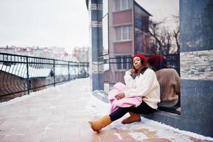 fille afro-américaine au chapeau rouge et manteau rose dans la rue de la ville contre la construction le jour de l'hiver. photo