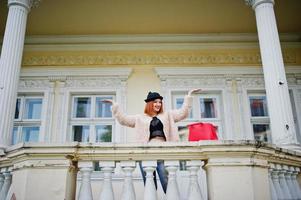 fille aux cheveux rouges dans des verres et un chapeau avec un sac à main rouge posé près de la maison vintage. photo