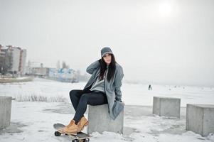 fille brune élégante en casquette grise, style de rue décontracté avec planche à roulettes le jour de l'hiver. photo