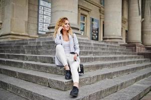 fille modèle blonde bouclée élégante porter sur blanc avec une tasse de café posant sur les escaliers en plein air. photo