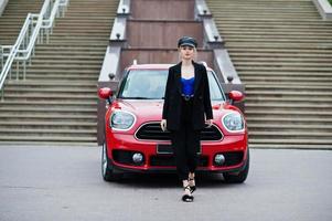 portrait d'une belle mannequin blonde sexy en casquette et en corset noir et bleu, avec un maquillage lumineux près d'une voiture de ville rouge. photo