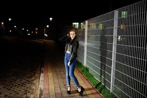 portrait de nuit d'une fille modèle portant un jean et une veste en cuir contre une clôture en fer. photo