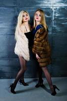 deux filles blondes élégantes portent un manteau de fourrure et une robe combinée posées contre un mur d'acier en studio. photo