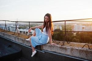 portrait d'une superbe jeune femme en t-shirt blanc et jupe bleue assise sur le bord de l'immeuble au coucher du soleil. photo