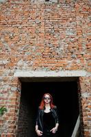 fille élégante aux cheveux rouges portant des lunettes de soleil en noir, contre un endroit abandonné avec des murs de briques. photo