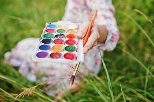 photo en gros plan de mains féminines tenant des peintures à l'aquarelle et un pinceau tout en peignant dans la nature.